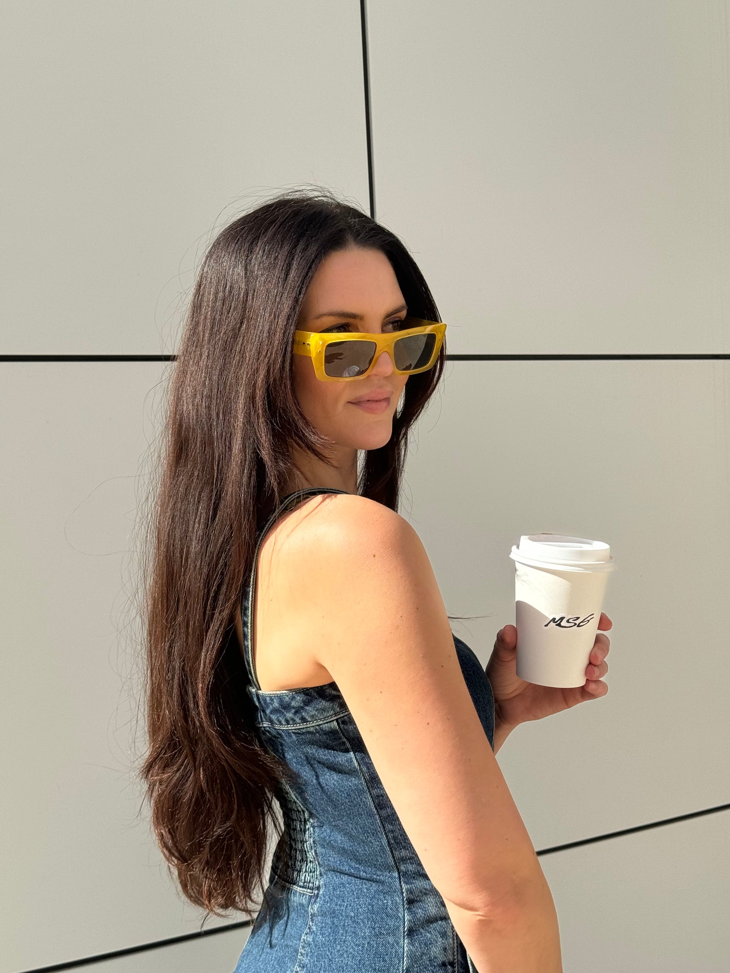 Woman holding a coffee cup with yellow sunglasses on, standing against a light-colored wall.